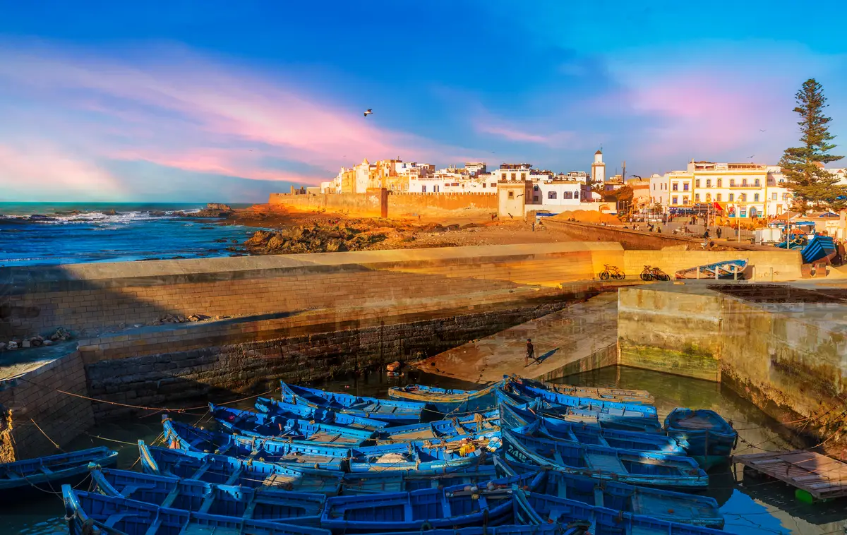The Essaouira Medina harbour featuring small blue fishing boats in the foreground, historic ramparts and buildings in the centre, and the Atlantic sea to the left. A popular transfer destination from Marrakech. 