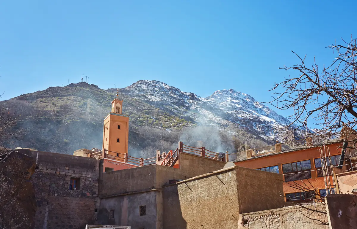 A scenic view of old buildings in the mountain village of Imlil, featuring a small mosque minaret and a nearby snowy peak of the High Atlas Mountains in the background.