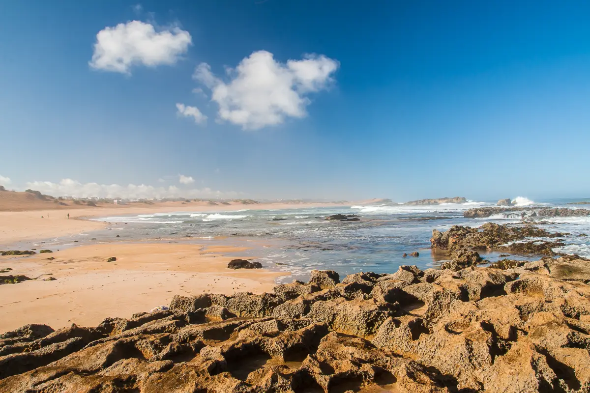 A wide sandy beach at Oualidia on a sunny day, featuring rocks in the foreground and a line of sand dunes sheltering beachfront properties.