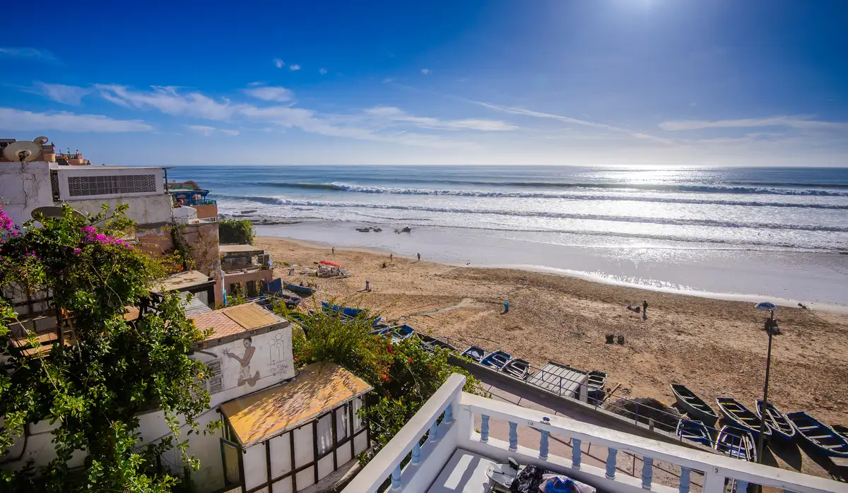 A sunny view of Taghazout Bay beach from a balcony, featuring rolling waves, sandy shores, and small beachfront properties.